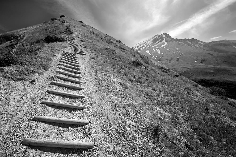 A black and white photo showing a staircase winding up a steep mountain trail with rugged peaks in the background.
