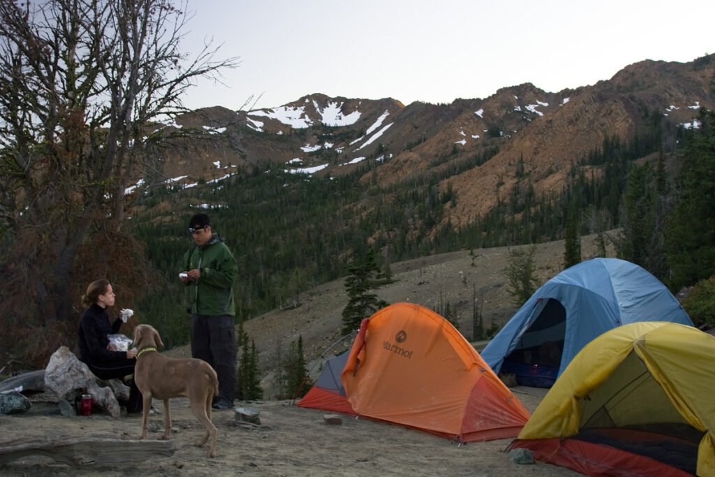 John and Kristie are camping near snow-capped mountains with their dog. Brightly colored tents stand out against the rugged landscape.