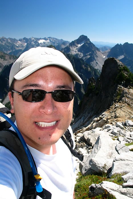 A happy hiker takes a selfie on a mountain trail under clear blue skies.