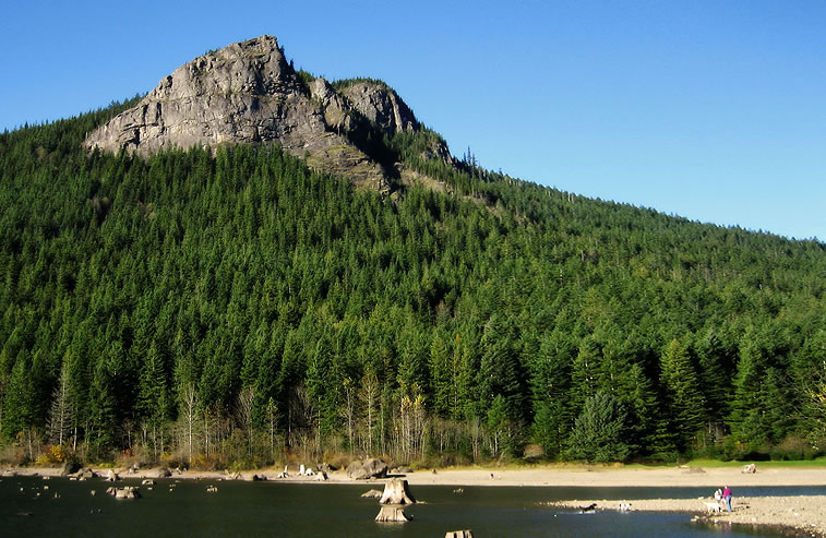 A scenic view of a mountain with dense green forest overlooking a calm lake. The clear blue sky enhances the natural beauty.