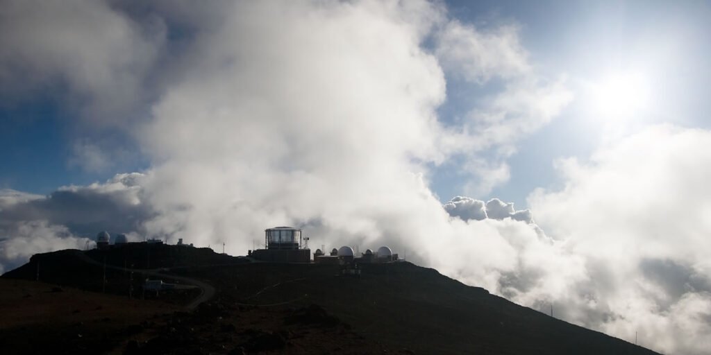 A panoramic view of a mountain with an observation deck at the summit, surrounded by dramatic clouds and a bright sun. The scene captures the grandeur of nature's beauty from an elevated viewpoint.