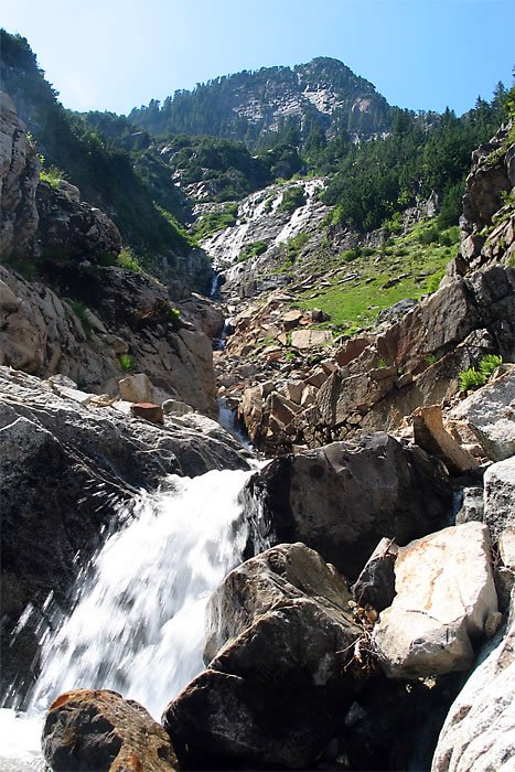 A cascading stream flows through rugged rocks, surrounded by lush greenery and towering mountains.