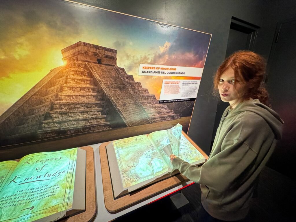 A person with red hair examines an open book titled 'Leaves of Knowledge' within a museum exhibit featuring a pyramid under a vibrant sunset sky.