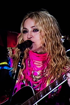 A woman performing with a guitar and singing into a microphone. Her outfit is vibrant pink with layered necklaces, complementing her curly blonde hair.