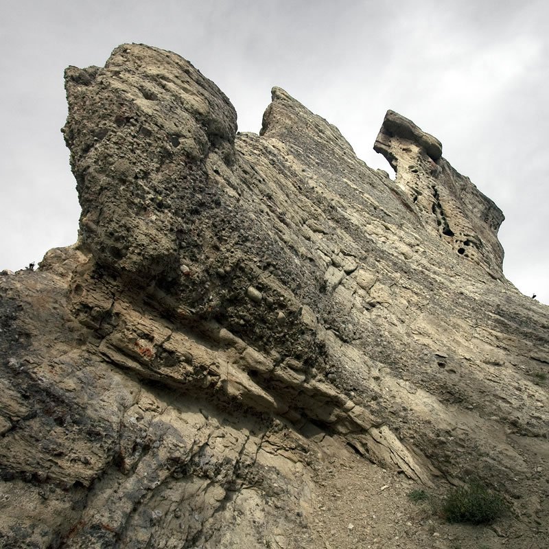A close-up view of an unusual rock formation with layered textures and sparse vegetation in the foreground. The sky is overcast, adding a dramatic backdrop to the rugged landscape.