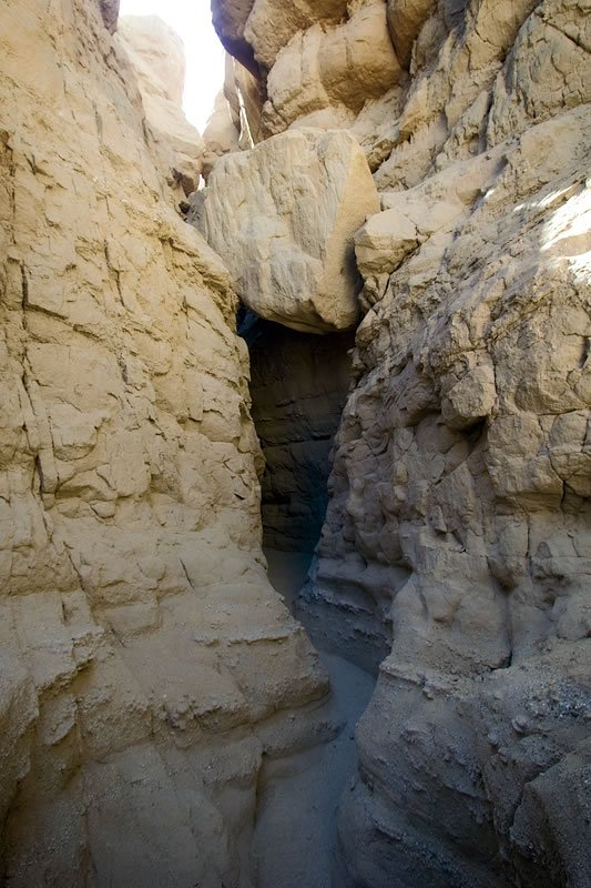 A narrow passage between towering, textured rock walls illuminated by sunlight from above.