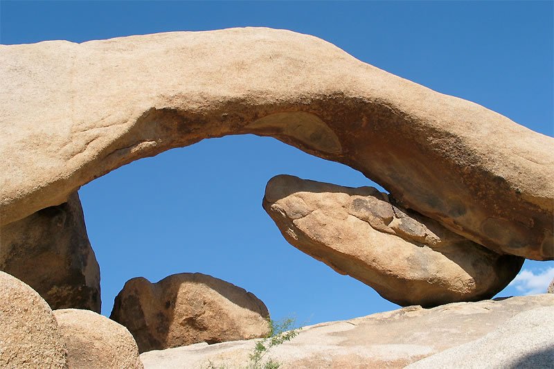 A massive natural rock arch against a clear blue sky, with smaller boulders surrounding the base. The archway provides a striking contrast to the texture and color of the sandstone.