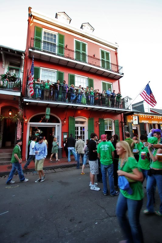 A bustling street in New Orleans filled with people wearing green shirts. The festive atmosphere is evident as the group gathers near vibrant buildings adorned with flags.