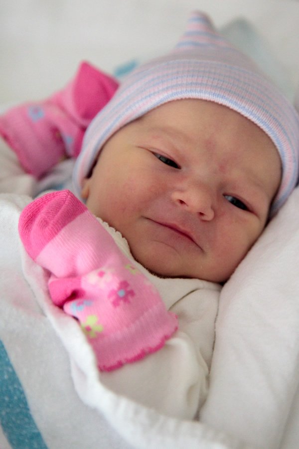 A newborn baby dressed in pink mittens and a striped beanie, lying on a soft blanket.