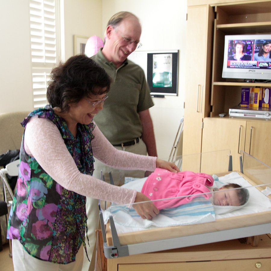 A woman and a man are smiling at a newborn baby lying in a hospital bassinet. The baby is wearing a pink outfit.