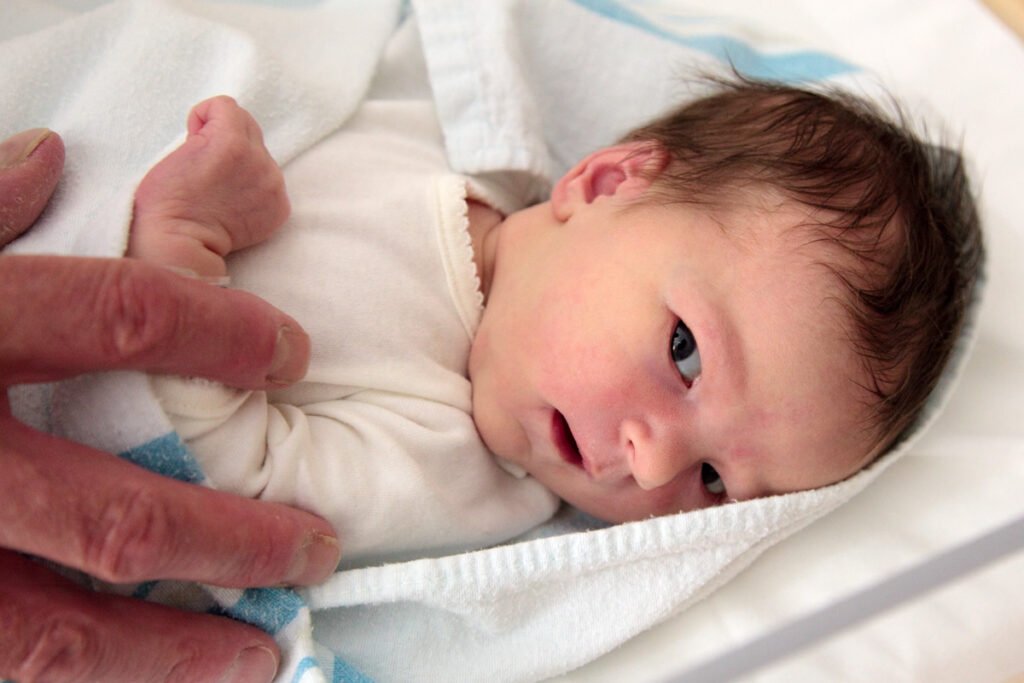 A newborn baby wrapped snugly in a white blanket, with tiny hands curled up. One small hand gently rests outside the blanket.
