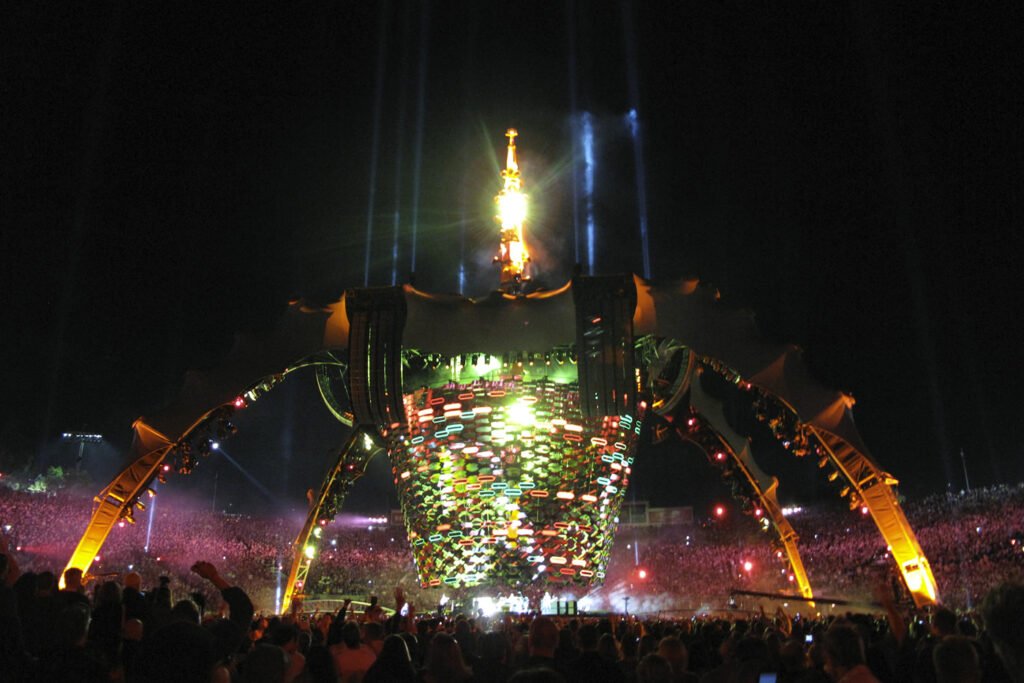 A large illuminated structure stands at the center of a nighttime festival. Crowds surround it under colorful lights.