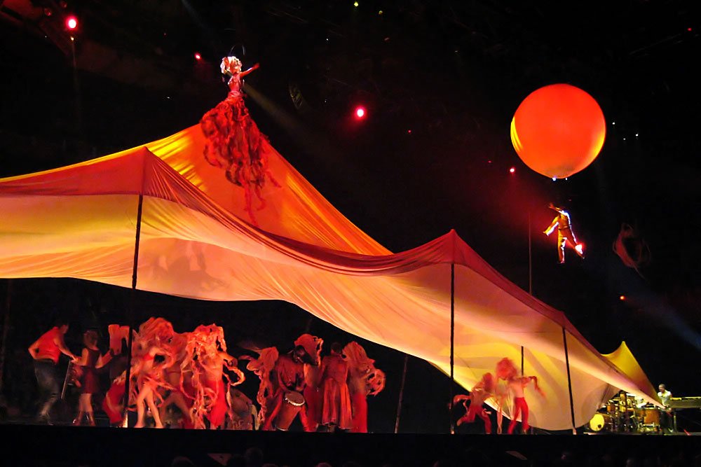 A bright red balloon floats above a glowing tent at night, with silhouettes of people underneath.