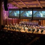 A live performance featuring an orchestra and choir on stage before a large audience.