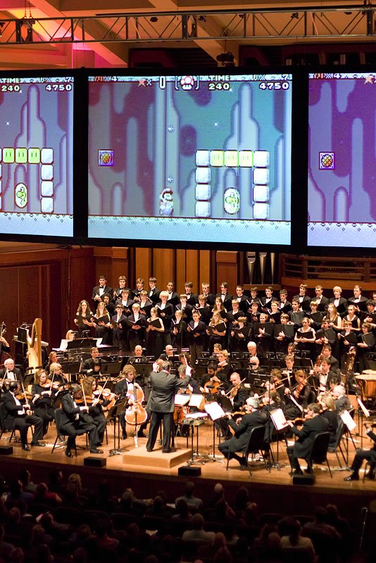 A full orchestra performs on stage below large projection screens. The screens display a retro-style video game with block-puzzle elements.