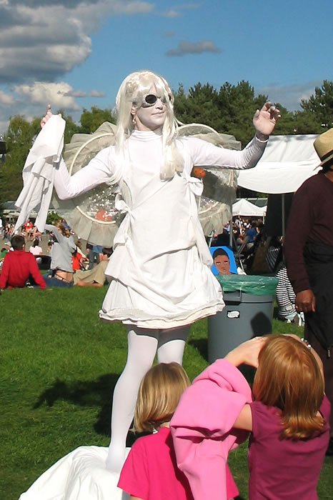 A model dressed in a white outfit with feathered wings performs at an outdoor event, captivating the audience. Spectators, including children in bright clothing, watch from the grass under a clear blue sky.