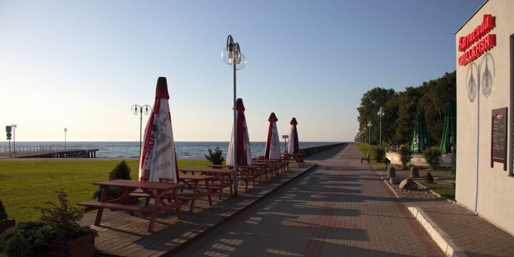 A scenic outdoor picnic area with wooden tables and umbrellas next to a paved walkway, overlooking the calm sea under a clear blue sky.