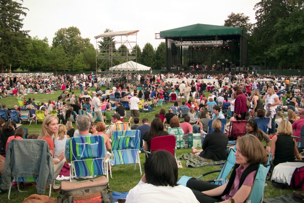A large crowd enjoys an outdoor concert under green trees with a stage setup.