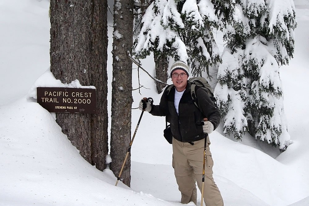 A hiker stands next to a Pacific Crest Trail sign in a snowy forest. The scene captures the essence of winter hiking in the wilderness.