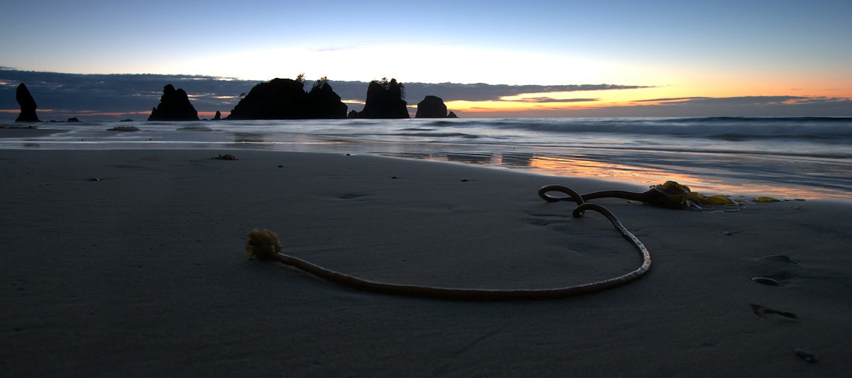 A serene beach scene featuring a coiled rope on the sand. In the background, sea stacks are silhouetted against a colorful sunset sky.