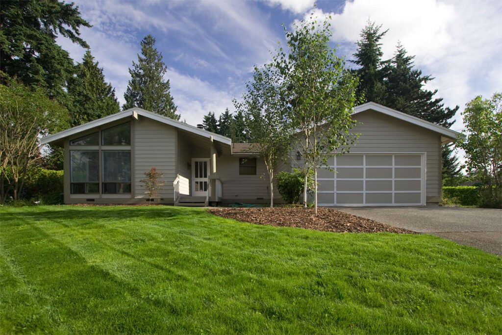 A charming single-story home in the Pacific Northwest, featuring a well-maintained green lawn, surrounded by tall trees and a clear blue sky.