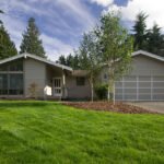 Painted Hills A charming single-story home in the Pacific Northwest, featuring a well-maintained green lawn, surrounded by tall trees and a clear blue sky.