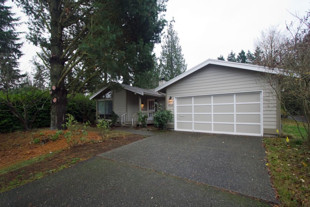 A white suburban home with a garage, surrounded by tall evergreen trees and a driveway, typical of a Pacific Northwest setting.