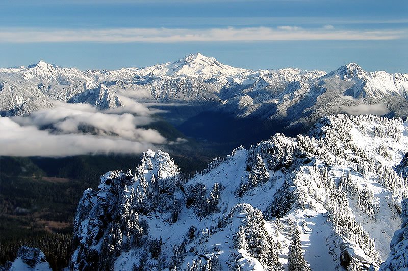 A stunning panoramic view of snow-capped mountains with clouds rolling through the valleys. The foreground features snow-dusted evergreen trees atop rocky outcrops, showcasing winter's beauty.
