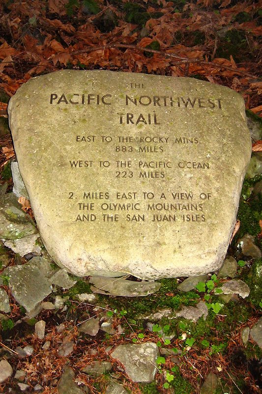 A carved stone marker on the Pacific Northwest Trail surrounded by forest floor foliage. The stone details distances to the Rocky Mountains, Pacific Ocean, and local view points.