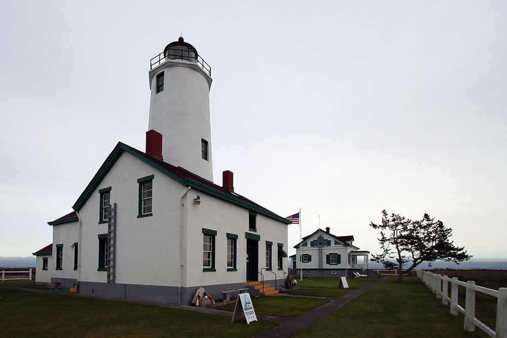A white lighthouse stands prominently on a hillside surrounded by a small building with a black roof. The scene is under a cloudy sky.