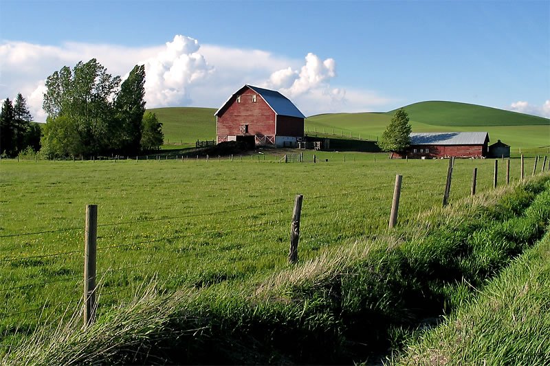 A picturesque red barn with a blue roof set against lush green hills under a clear sky.