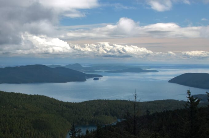 A breathtaking view of rugged coastal islands surrounded by vast blue waters, with lush green forests below under a partly cloudy sky.