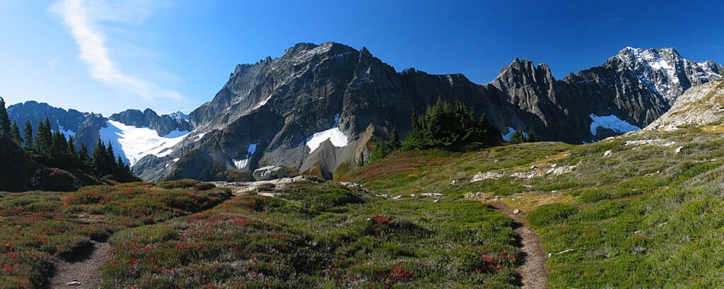 A scenic view of a hiking trail meandering through lush greenery with snow-capped mountains in the background under a clear blue sky.