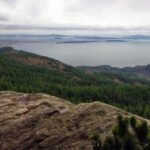 Dungeness Spit Panoramic view of the Pacific Northwest showcasing dense forests, expansive water bodies, and distant islands under a cloudy sky. A rugged rock formation is prominent in the foreground.