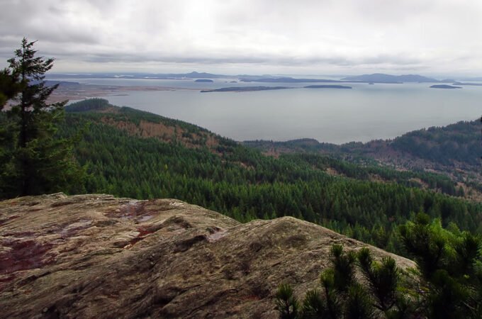 Panoramic view of the Pacific Northwest showcasing dense forests, expansive water bodies, and distant islands under a cloudy sky. A rugged rock formation is prominent in the foreground.