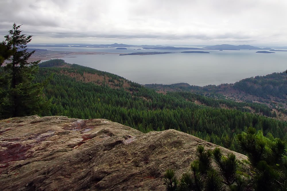 Panoramic view of the Pacific Northwest showcasing dense forests, expansive water bodies, and distant islands under a cloudy sky. A rugged rock formation is prominent in the foreground.