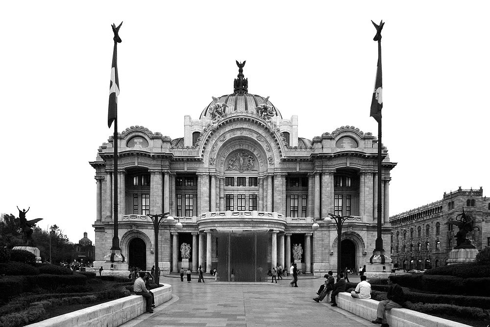 A grand historical building with ornate architecture and flags atop. People are seated on benches outside.
