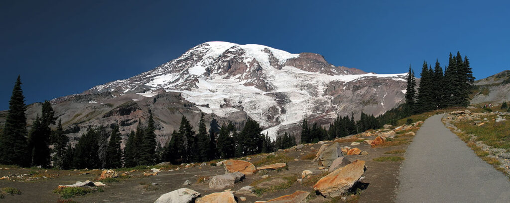 A panoramic view showing Mount Rainier's snow-covered peak and a trail leading into the distance.