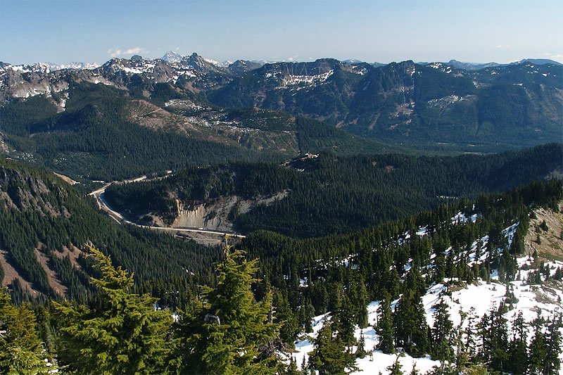 A breathtaking view of the Pacific Northwest mountains, showcasing snow-covered peaks and lush green forests with patches of lingering snow.