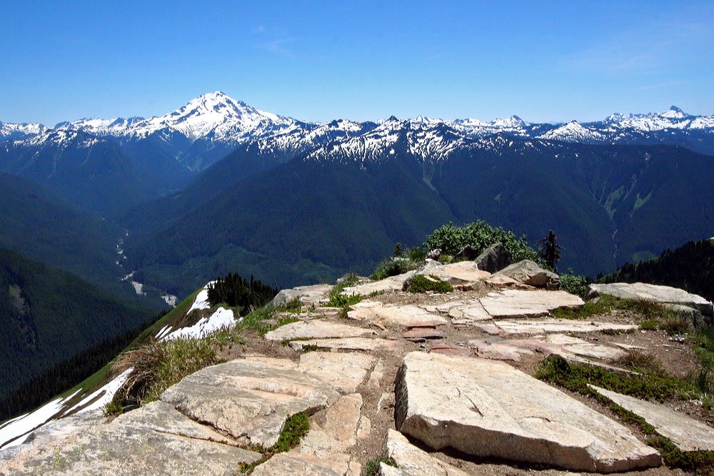 A stunning panoramic view of snow-capped mountains under a clear blue sky, with rocky terrain in the foreground.