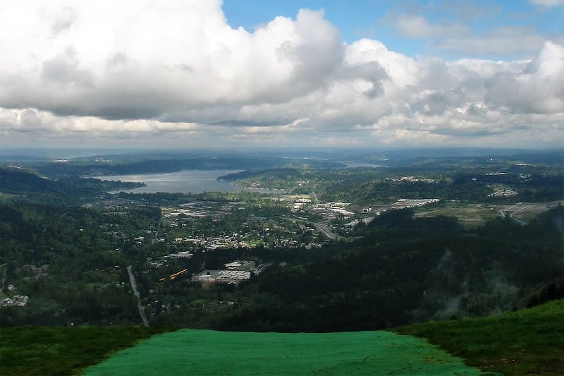 A breathtaking view from a high vantage point, showcasing a sprawling cityscape bordered by water and surrounded by lush green hills under a partly cloudy sky.