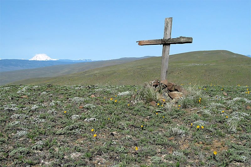 A wooden cross stands on a grassy hill with distant mountains and a clear blue sky.