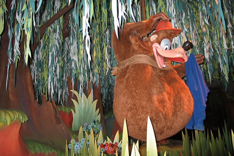 A character from the movie Peter Pan, resembling a Lost Boy, stands amidst lush greenery and hanging vines in a whimsical forest setting. The figure wears a cap and holds a flute, ready to play.