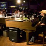 PLAY! A musician plays piano under warm stage lighting in a dimly lit venue.