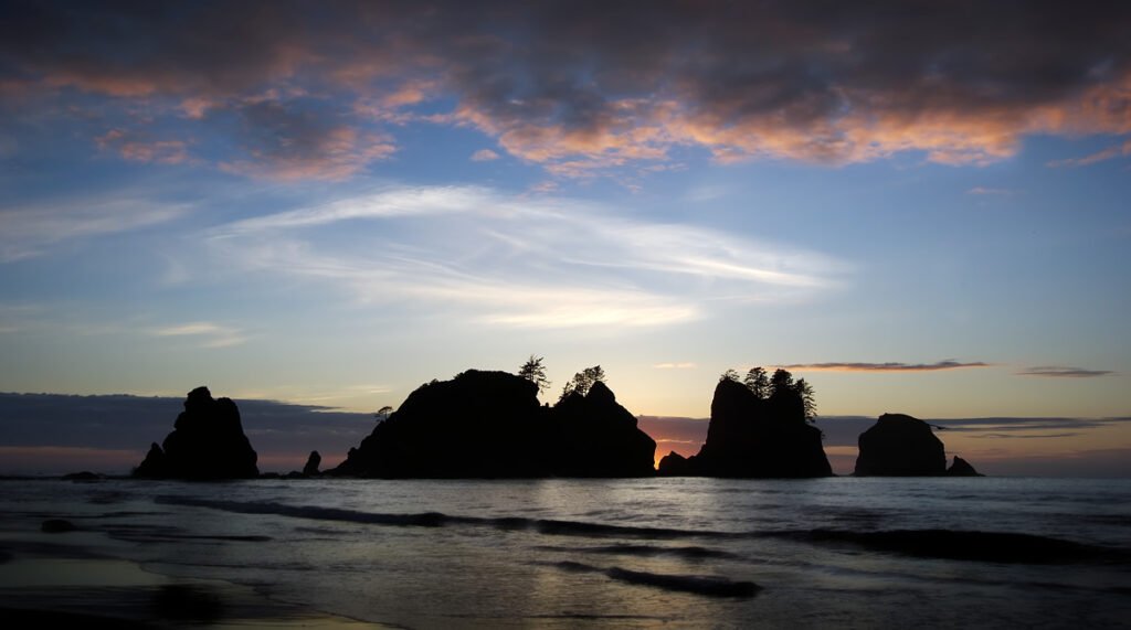 Silhouetted sea stacks stand against a colorful sunset sky over calm ocean waters.