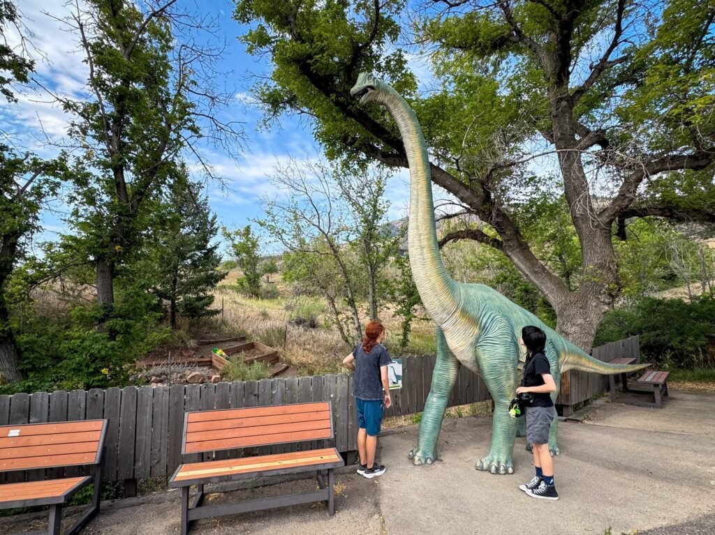 Two individuals standing next to a large dinosaur statue in a lush park setting.