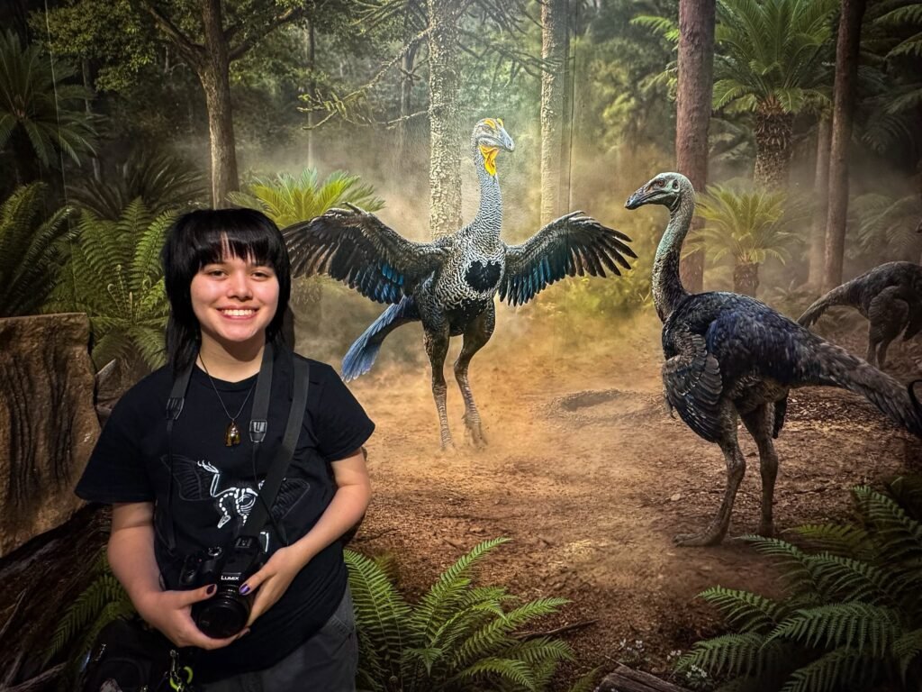 Person holding a camera poses in front of a vibrant fern and prehistoric bird display.