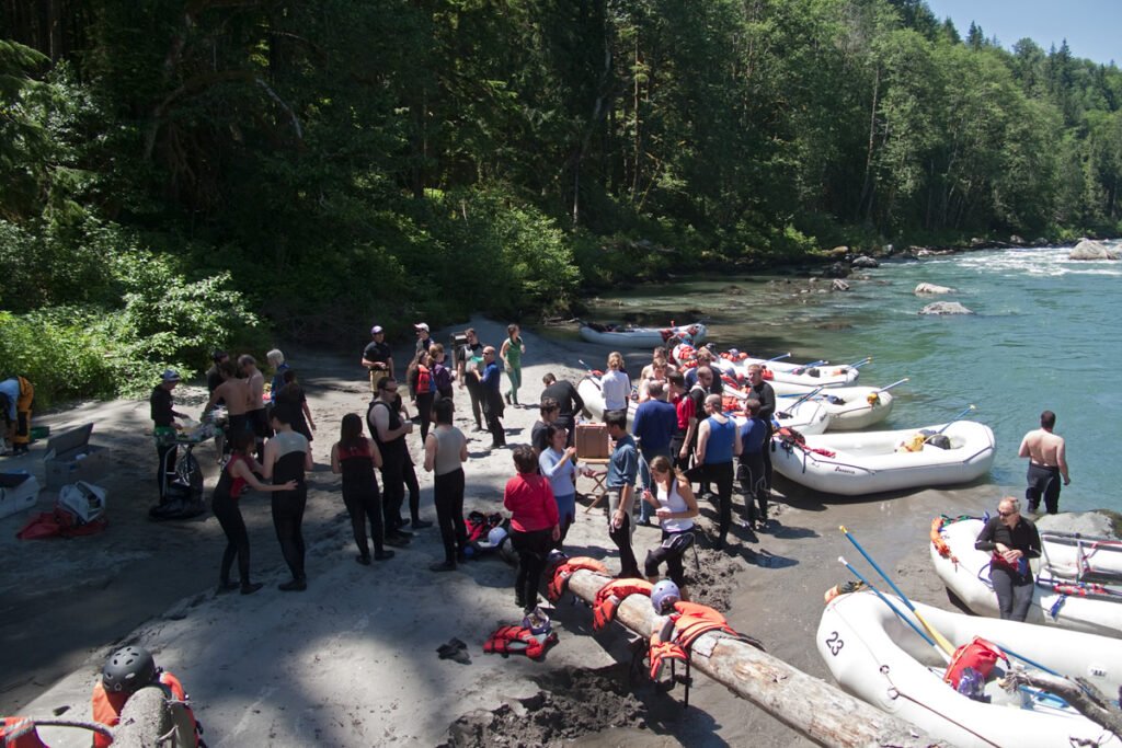 A diverse group of people gathered on a sandy riverside, preparing white inflatable rafts with paddles and life jackets, amidst a lush green forest backdrop.