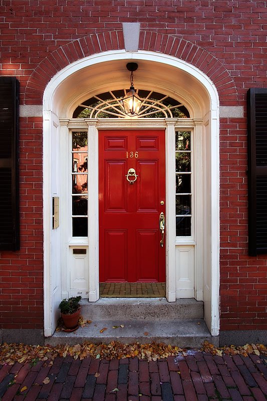 A bright red front door framed by a white archway and flanked by sidelights, set against a red brick wall with fallen leaves on the ground.