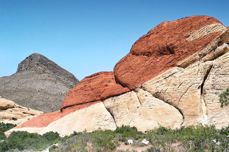 A striking view of red and cream-colored rock formations with green shrubs in the foreground under a clear blue sky. The layered rock formations create a unique and vibrant desert landscape.